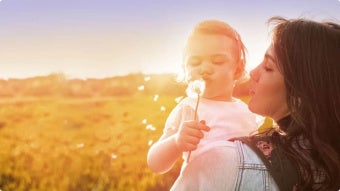 Junge Frau hält ein Kind, dass auf einen Löwenzahn pustet für den Beba Expert HA 2 & Beba Expert HA 3 Produkttest