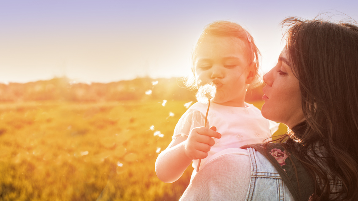 Junge Frau hält ein Kind, dass auf einen Löwenzahn pustet für den Beba Expert HA 2 &amp; Beba Expert HA 3 Produkttest